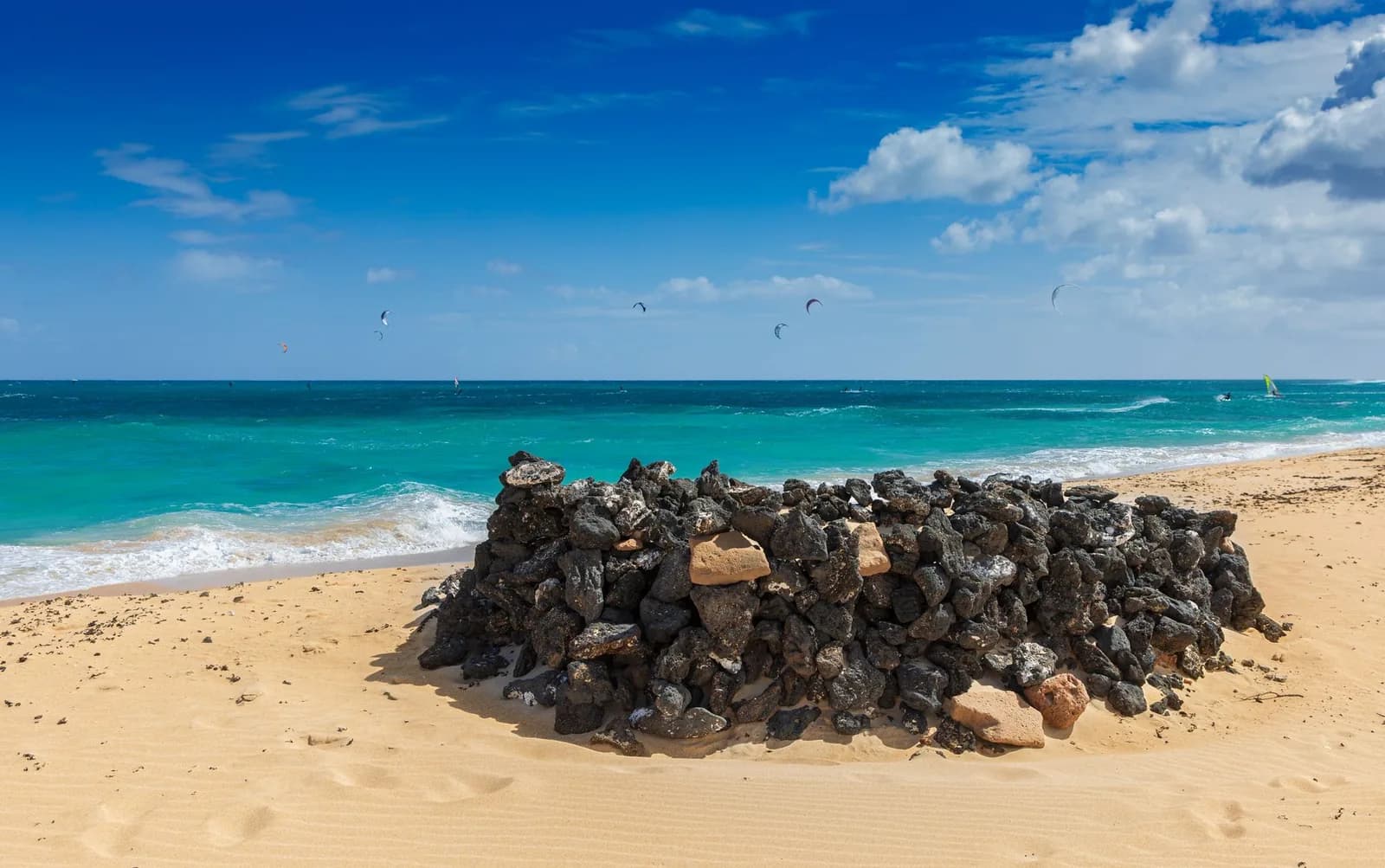 Plage de sable et abri en pierres volcaniques à Corralejo, kitesurfeurs au loin sur l'eau turquoise, Fuerteventura
