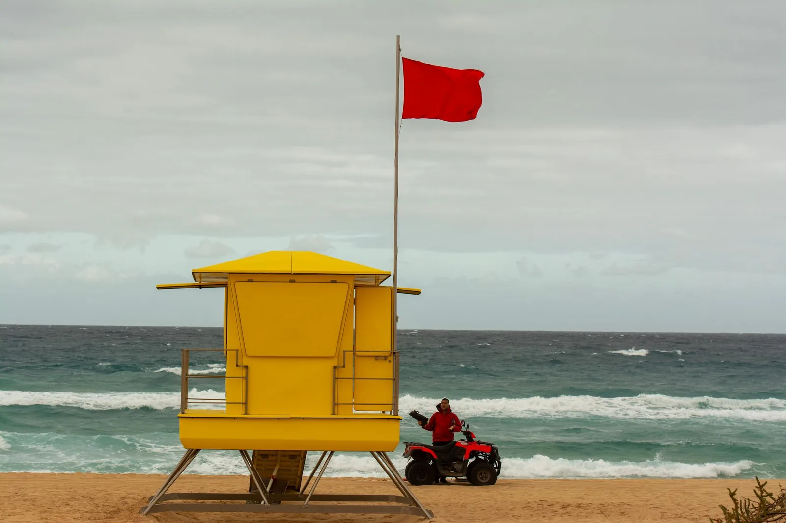 Sauveteur et son quad au pied d'un poste de secours jaune, drapeau rouge hissé face à une mer agitée, Canaries